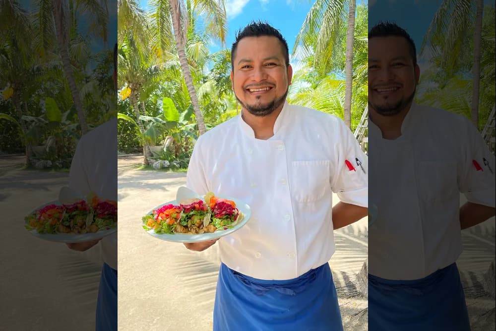 Smiling man in a white chef’s jacket and blue apron presenting a white platter filled with tacos topped with colorful vegetables