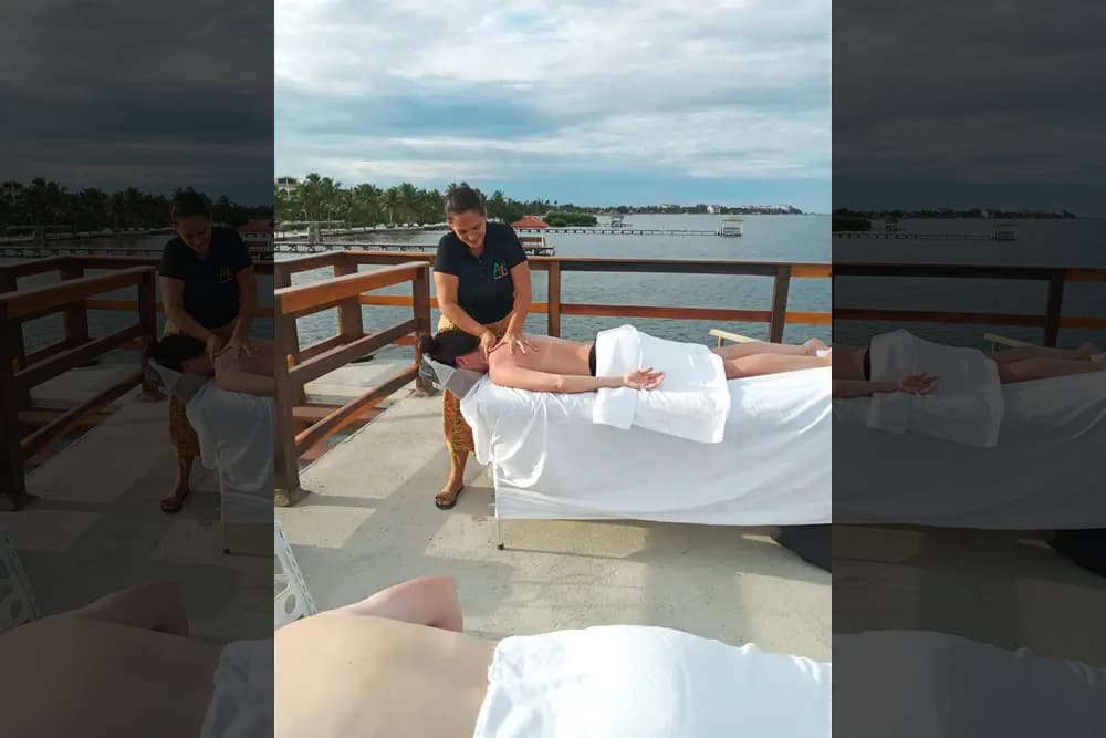 Masseuse rubbing back and shoulders of person lying face down on massage table draped in white towels on deck overlooking ocean